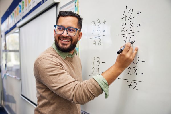 Happy teacher teaching how to do the column additions at elementary school. Smiling man explaining additions in column in primary class while writing on whiteboard. Math’s teacher explaining arithmetic sums to elementary children.